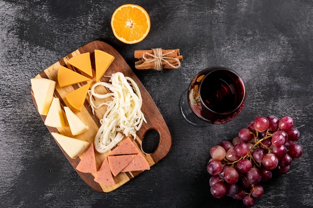 Vista superior de vino tinto con uva, naranja y queso sobre tabla de cortar de madera sobre una superficie oscura horizontal