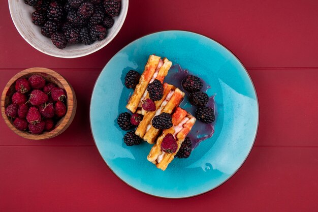 Vista superior de waffles dulces con frambuesas y moras en una placa azul sobre una mesa roja