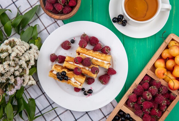 Vista superior de waffles dulces con frambuesas en un plato con cerezas blancas y flores sobre una toalla a cuadros