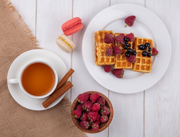 Vista superior de waffles dulces en un plato con una taza de té, canela y frambuesas con macarrones sobre una mesa blanca