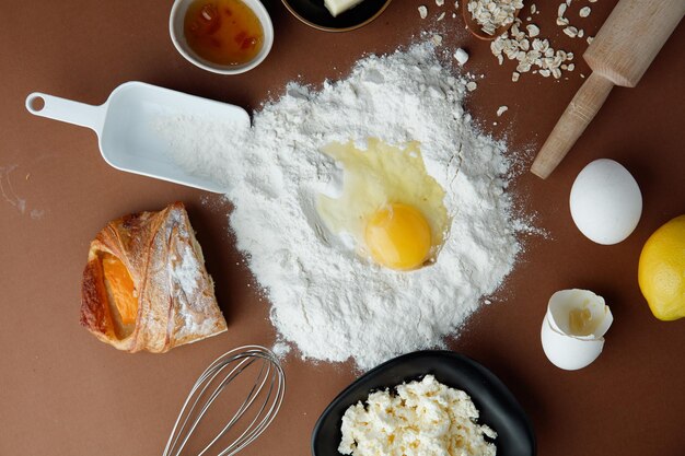 Vista superior de la yema de huevo en harina con mermelada, croissant, medio requesón, limón, huevo, avena con cuchara de medida, rodillo y batidor sobre fondo blanco.