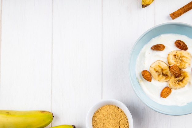 Vista superior de yogurt con plátano y almendras, palitos de canela en blanco con espacio de copia