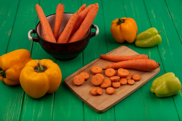 Foto gratuita vista superior de zanahorias vegetales de raíz en un recipiente con rodajas picadas en una tabla de cocina de madera con pimientos aislados en una pared de madera verde