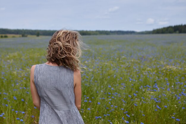 Vista trasera de una chica esbelta irreconocible que disfruta de un hermoso paisaje, de pie en medio de un prado verde con flores azules, su cabello rubio rizado ondeando en el viento. Mujer caminando al aire libre