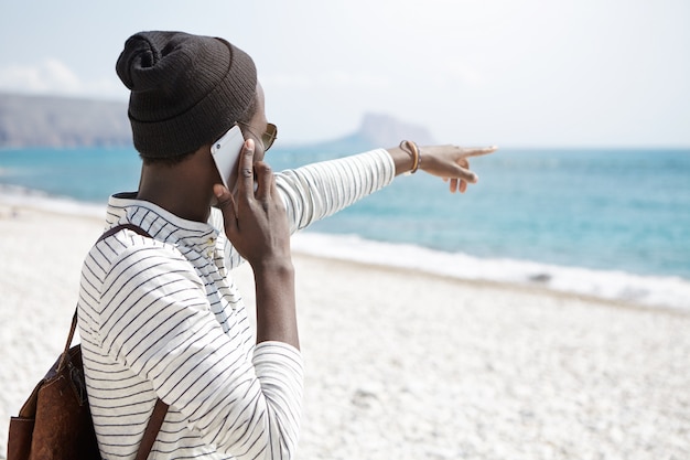 Vista trasera del elegante hombre afroamericano que señala el dedo hacia el océano mientras está de pie en la playa y hablando por teléfono móvil, notando algo interesante en el agua. Gente y tecnología moderna