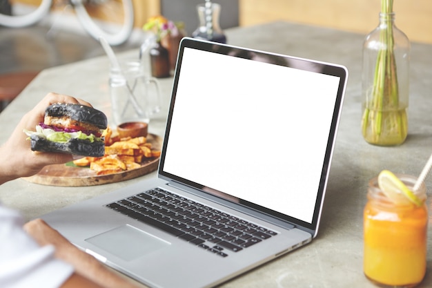 Foto gratuita vista trasera del estudiante sentado delante de una computadora portátil genérica abierta con una hamburguesa en la mano