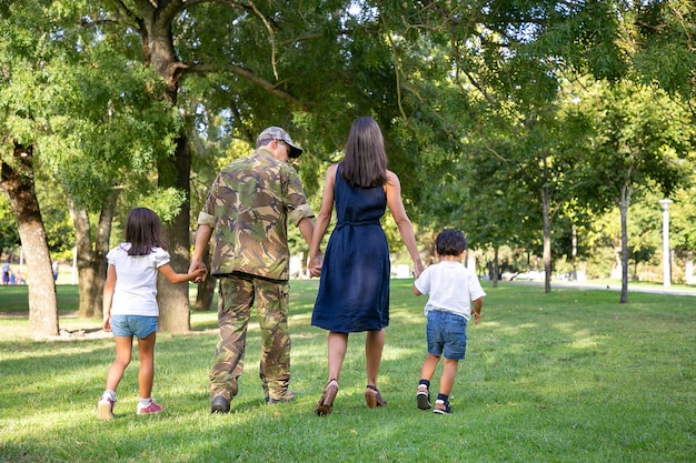 Vista trasera de la familia caucásica tomados de la mano y caminar juntos en el parque de la ciudad. Papá con uniforme de camuflaje, mamá de pelo largo y niños disfrutando de las vacaciones en la naturaleza. Concepto de reunión familiar y fin de semana.