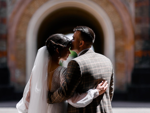 Vista trasera de una hermosa pareja vestida con trajes de boda abrazándose unos a otros mientras posan en el fondo del edificio del arco durante el día soleado