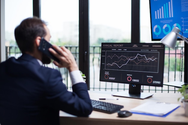 Vista trasera del joven empresario hablando por teléfono con su socio comercial. Gráficos digitales en el monitor de la computadora.