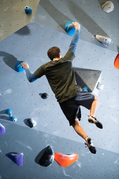 Vista trasera de un joven haciendo ejercicio de escalada en el gimnasio. Atleta caucásico en ropa deportiva haciendo boulder sosteniendo rocas artificiales en la pared con manos fuertes y colgando en el aire. Deporte extremo y vida sana