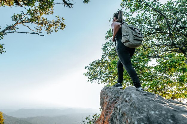 Vista trasera de una joven mujer de senderismo asiática de pie en el punto de vista y luciendo una hermosa vista con feliz en la montaña máxima y espacio de copia de rayos solares