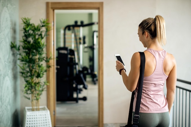 Vista trasera de una mujer atlética enviando mensajes de texto por teléfono en un pasillo del gimnasio