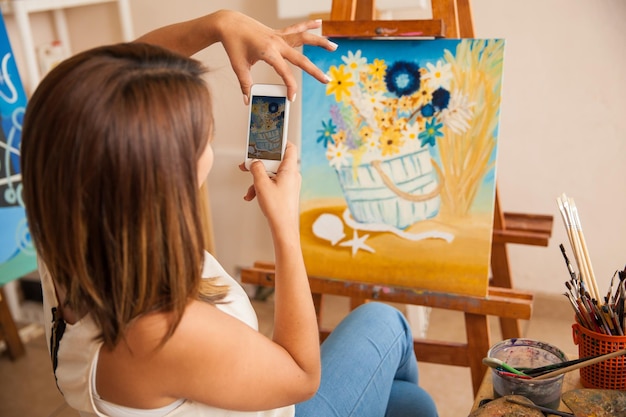 Vista trasera de una mujer joven tomando una foto de su pintura más reciente para compartirla y venderla en línea