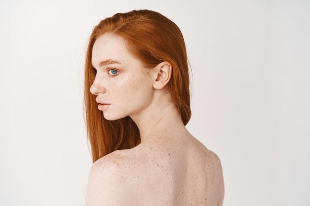 Vista trasera de la mujer pelirroja hacia atrás, gira la cabeza y mira a la izquierda soñadora. Modelo de mujer pálida con el pelo rojo posando en la pared blanca