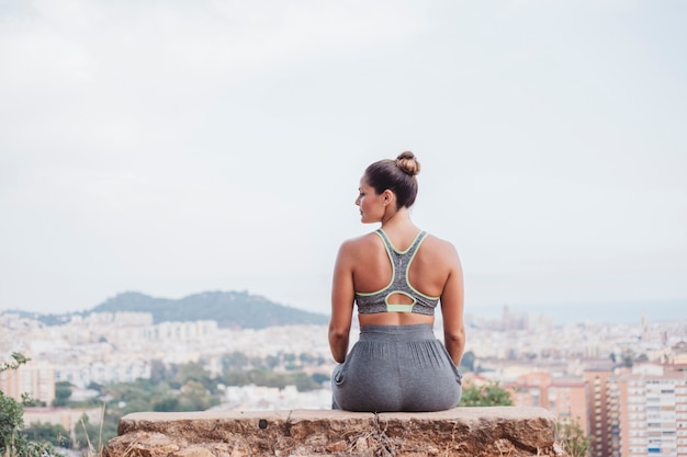 Vista trasera de mujer sentada en roca enfrente de ciudad