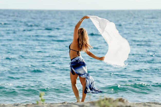 Vista trasera de la mujer sintiéndose libre mientras sostiene un chal en un día ventoso junto al mar