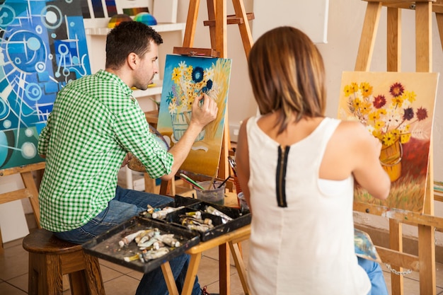 Vista trasera de un par de adultos jóvenes que trabajan en sus propias pinturas mientras estudian en una escuela de arte