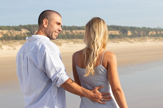 Vista trasera de una pareja cariñosa caminando por la playa. Hombre caucásico con cabeza rapada y mujer vestida de forma casual caminando, abrazándose en la cintura. Amor, vacaciones, concepto de afecto.