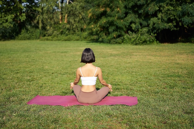 Foto gratuita vista trasera de la silueta de la mujer haciendo yoga sentada en la alfombra de fitness y meditando en el césped verde