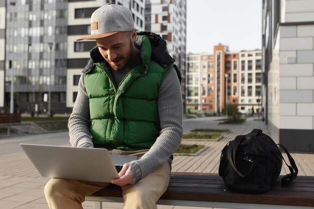 Vista urbana de un hombre joven sin afeitar que lee noticias, revisa el correo electrónico o escribe un mensaje en línea usando una computadora portátil al aire libre, sentado en un banco con una bolsa, viajando solo, disfrutando de wifi gratis