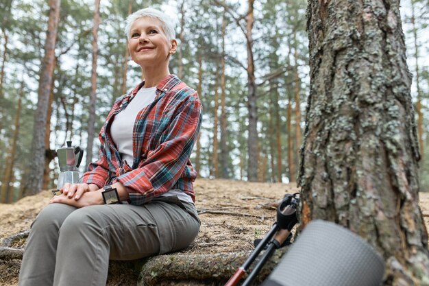 Vista de verano al aire libre de una atractiva mujer de mediana edad aventurera sentada junto al árbol, hirviendo agua para el té en la tetera, con una mirada alegre, admirando la hermosa naturaleza, pájaros cantando, sonriendo feliz