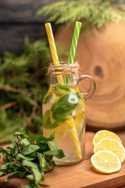 Vista vertical de agua de desintoxicación fresca en un vaso servido con tubos y limones sobre una tabla de cortar de madera sobre una mesa marrón