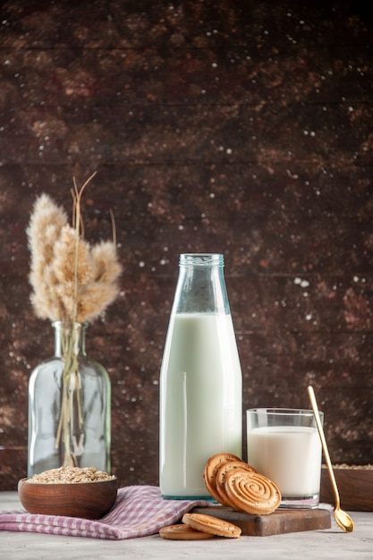 Vista vertical de la botella de vidrio abierta y la taza llena de galletas de leche, avena en una olla marrón sobre una toalla despojada de color púrpura en la tabla de cortar de madera