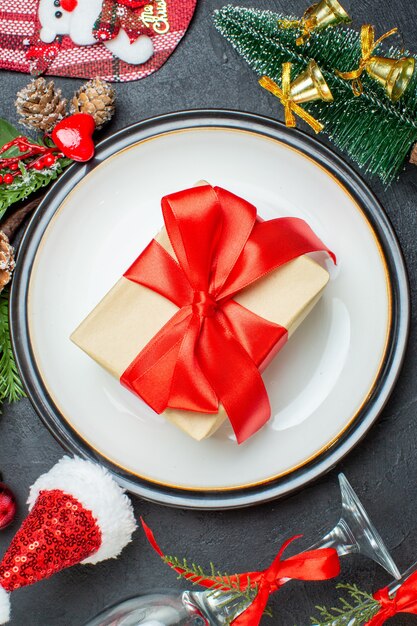 Vista vertical de la caja de regalo en el plato de cena árbol de navidad ramas de abeto cono de coníferas sombrero de santa claus copas de vidrio caído sobre fondo negro