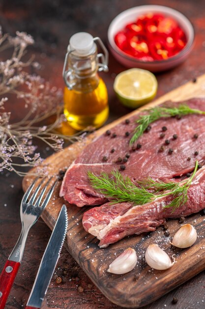 Vista vertical de la carne roja en la tabla de cortar de madera y el ajo pimiento verde tenedor y cuchillo sobre fondo oscuro