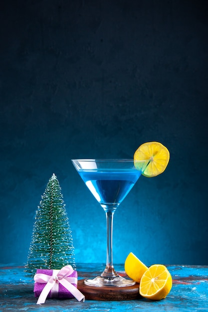 Vista vertical del cóctel alchocol en una copa de vidrio servido con una rodaja de limón y caja de regalo árbol de navidad sobre fondo azul.