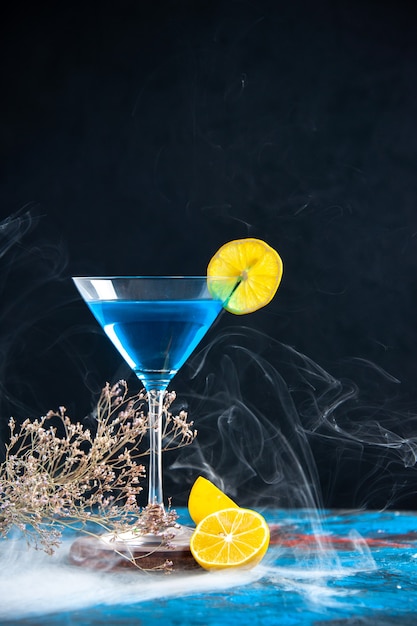 Vista vertical del cóctel de alchocol en una copa de vidrio servido con una rodaja de limón y ramas de abeto humo sobre una mesa azul
