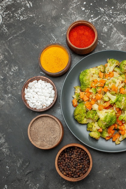 Vista vertical de comida saludable con brocoli y zanahorias en un plato negro y especias en mesa gris