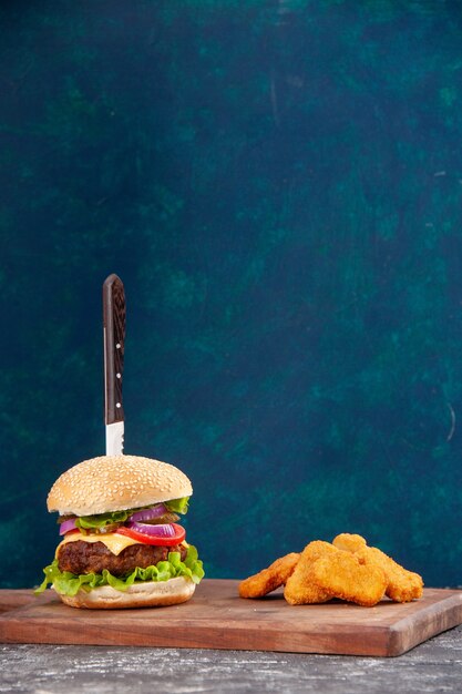 Vista vertical de cuchillo en sabroso sándwich de carne y nuggets de pollo sobre tabla de madera sobre superficie azul oscuro