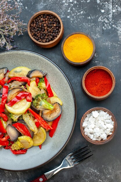 Vista vertical de una deliciosa comida vegetariana en un plato y tenedor junto a diferentes especias sobre fondo oscuro