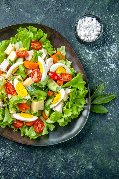 Vista vertical de deliciosa ensalada casera con muchos ingredientes en un plato y sal sobre fondo de colores mezcla verde negro