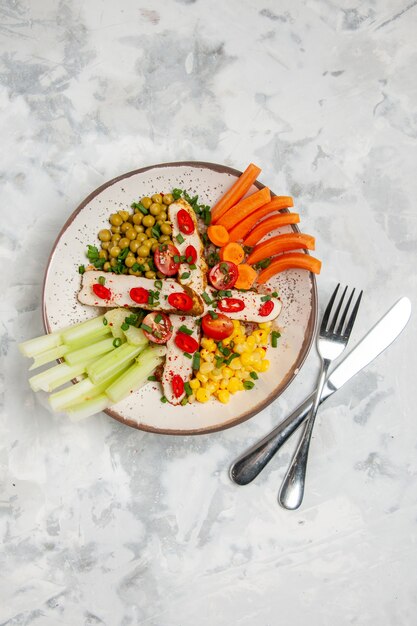 Vista vertical de una deliciosa ensalada con varios ingredientes en un plato y cubiertos en superficie blanca