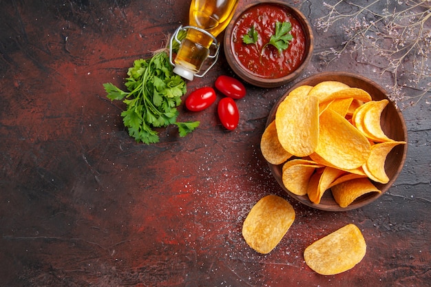 Vista vertical de deliciosas patatas fritas crujientes caseras en un pequeño tazón marrón botella de aceite caído verde y tomates ketchup de ajo en el cuadro oscuro