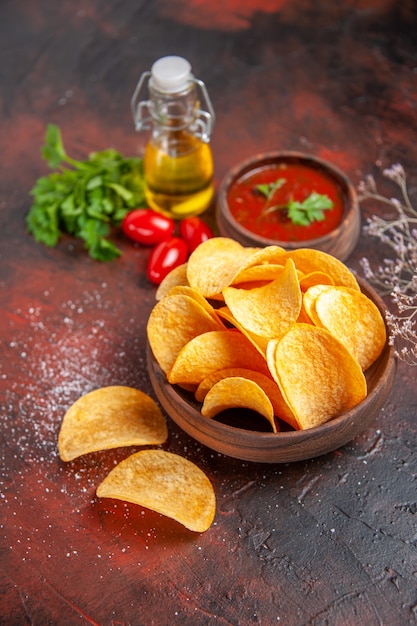 Vista vertical de deliciosas patatas fritas crujientes caseras en un pequeño tazón marrón botella de aceite tomates verdes ketchup de ajo en el cuadro oscuro