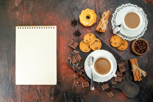 Vista vertical de un delicioso café en tazas blancas en la tabla de cortar de madera, galletas, canela, limas, barras de chocolate, cuaderno de espiral sobre fondo de colores mezclados