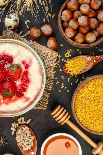 Vista vertical del delicioso desayuno servido con mermelada de fresas en un bol y cucharas de avellanas de miel sobre fondo de color oscuro