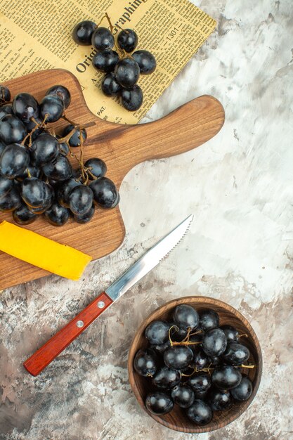Vista vertical del delicioso racimo de uva negra fresca y queso sobre una tabla de cortar de madera y en un cuchillo marrón sobre fondo de colores mezclados