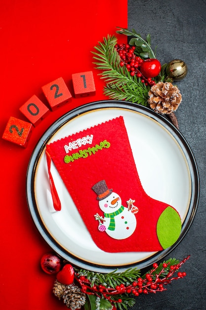 Vista vertical del fondo de año nuevo con calcetín de navidad en el plato de cena accesorios de decoración ramas de abeto y números en una servilleta roja sobre una mesa negra