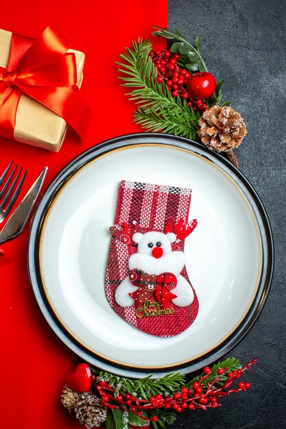 Vista vertical de fondo de año nuevo con calcetín de Navidad en plato cubiertos accesorios de decoración ramas de abeto junto a un regalo en una servilleta roja