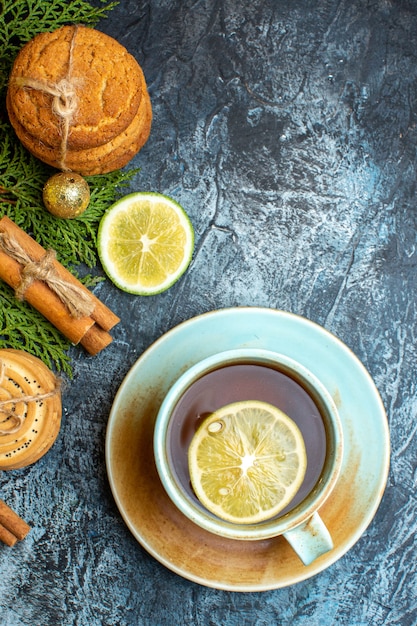 Vista vertical de galletas apiladas y ramas de abeto limón canela limas junto a una taza de té negro sobre fondo oscuro