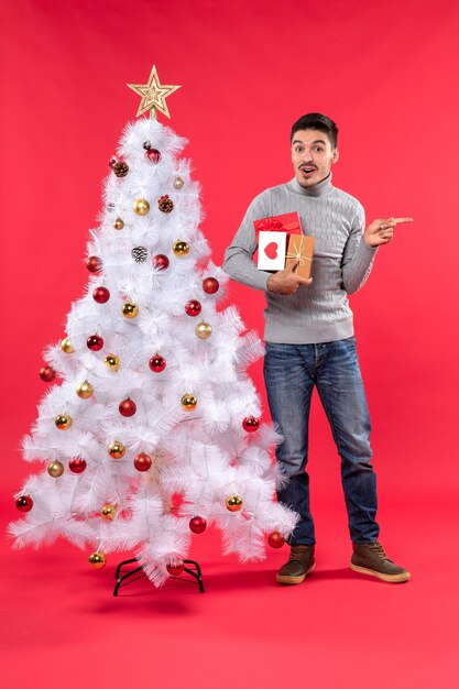 Vista vertical del hombre guapo confundido de pie cerca del árbol de Navidad blanco decorado y sosteniendo sus regalos