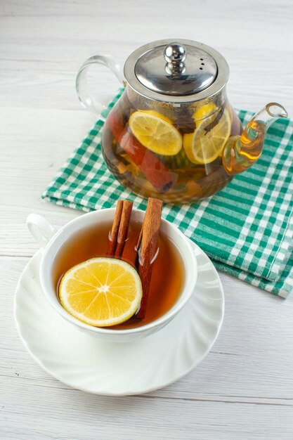 Vista vertical de la hora del té con té de hierbas mezclado con limón en una olla de vidrio y una taza sobre una toalla pelada verde sobre fondo blanco.