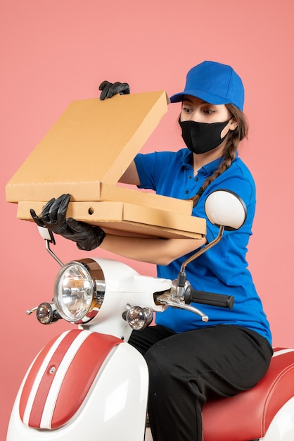 Vista vertical del joven mensajero confía en llevar máscara médica y guantes abriendo cajas en melocotón pastel