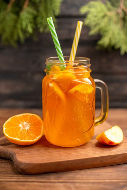 Vista vertical de jugo de fruta fresca en un vaso servido con tubos y manzana y naranja sobre una tabla de cortar de madera sobre una mesa marrón