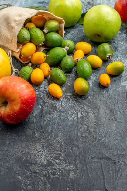 Vista vertical de kumquats frescos dentro y fuera de una pequeña bolsa blanca caído y manzanas verdes amarillas rojas en el cuadro gris