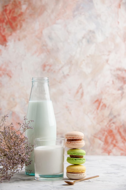 Vista vertical de la leche fresca en una taza de vidrio y una botella junto a las coloridas y deliciosas flores de macarons cuchara dorada en el lado derecho sobre fondo de colores pastel